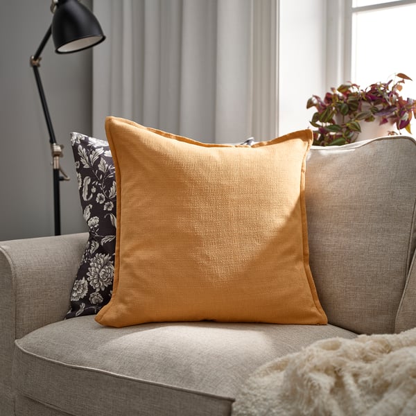 Salon confortable avec coussin LAGERPOPPEL jaune sur un canapé gris, contrastant un coussin à motifs floraux et un lampadaire noir.