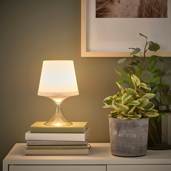 Petite lampe de table avec abat-jour blanc posée sur une pile de livres. À côté d’une plante en pot dans une jardinière en béton, contre un mur avec une photo encadrée.
