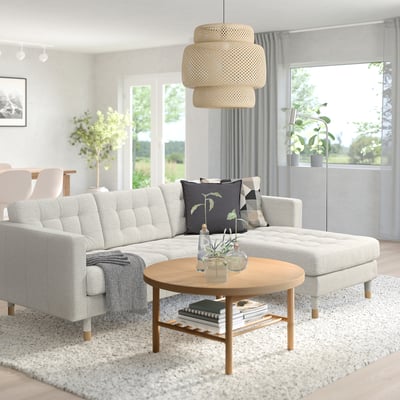 Salon moderne : canapé en forme de L landskröna gris, table basse en bois naturel, coussins floraux, lampe texturée suspendue, grandes fenêtres, plantes d’intérieur.