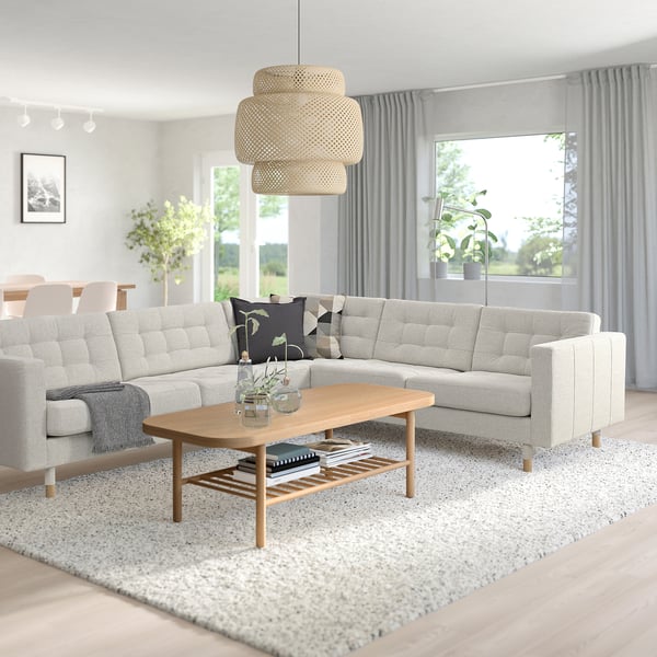 Salon moderne avec canapé en forme de L gris clair, table basse en bois et suspension. Des rideaux gris encadrent les fenêtres, laissant entrer la lumière naturelle.