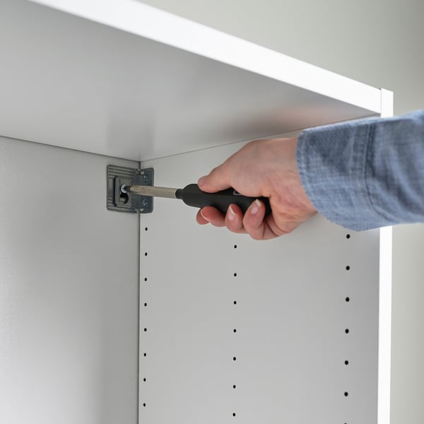Une personne assemble un meuble blanc avec un tournevis, qu’il fixe au mur. L’armoire a des trous visibles pour les vis.