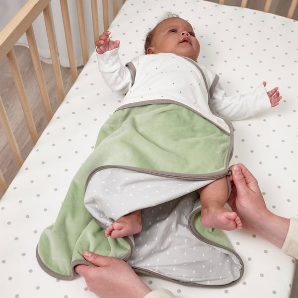 Bébé dans une gigoteuse verte, zippée, dans un lit bébé en train d’être habillé.