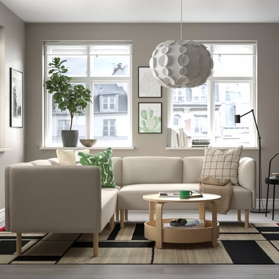 Salon moderne avec un canapé beige en forme de L, une table basse en bois, une grande fenêtre, une lampe suspendue, une plante et un tapis.