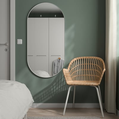 Chambre moderne : mur vert, grand miroir ovale blanc, chaise en osier, rideaux blancs, lit. Miroir plus grand que la chaise.