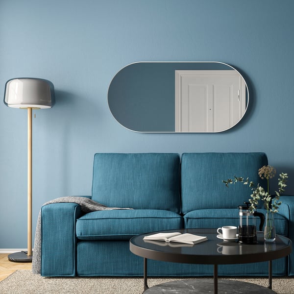 Un miroir ovale blanc LINDBYN au-dessus d’un canapé bleu, porte réfléchissante et lampe.