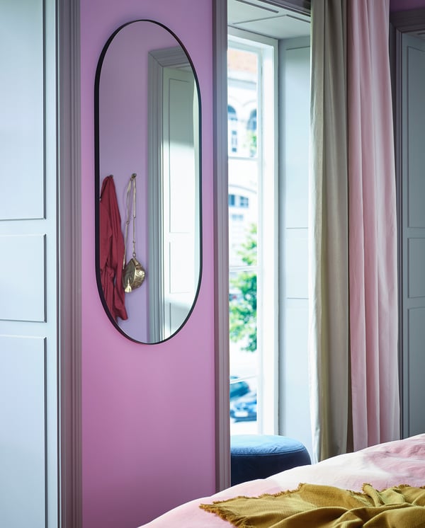 Chambre moderne avec miroir LINDBYN reflétant les accessoires rouges sur le mur lavande, rideaux roses, lumière naturelle.