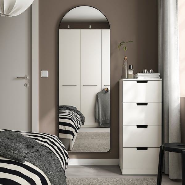 Chambre moderne avec miroir arqué LINDBYN, grande armoire blanche, lit à cadre noir, tiroirs blancs et tabouret. Le miroir et l’armoire dominent l’espace.