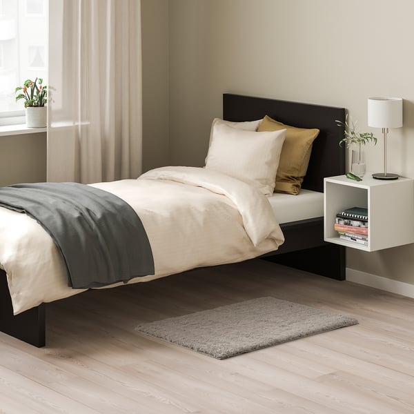 Chambre avec tapis beige LINDKNUD, lit blanc, tête de lit noire et table de chevet avec lampe. Le tapis est petit, s’adaptant sous le lit.