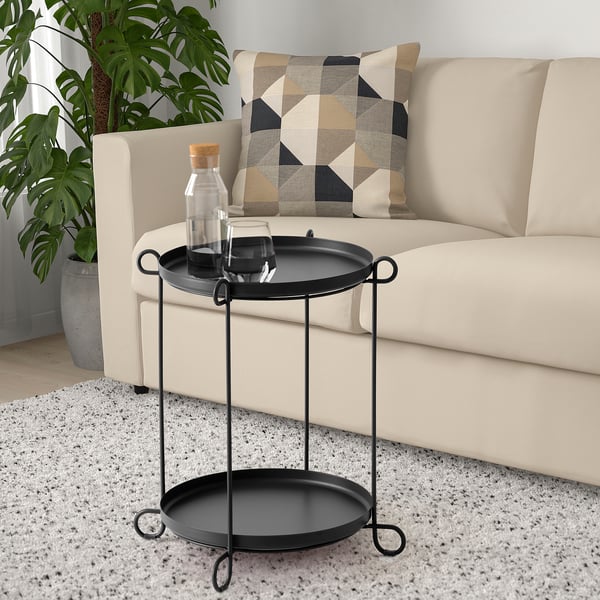 Table d’appoint noire avec deux plateaux ronds et poignées en boucle, s’adaptant à côté du canapé et près d’une grande plante en pot.