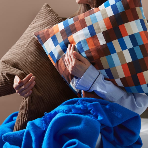 Personne tenant un coussin LÖVNATURSKOG avec fermeture éclair, carreaux multicolores, sur un coussin beige et une couverture bleue.