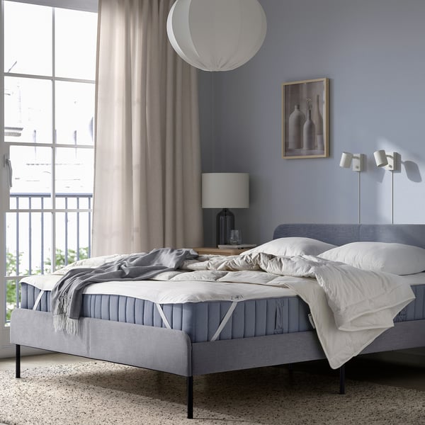 Chambre moderne avec protège-matelas LUDDROS gris, oreillers, rideaux et vue sur la ville.