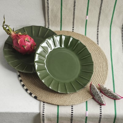 Une assiette en grès MÄVINN vert repose sur un napperon rond tissé avec des fruits du dragon. Le bord festonné des assiettes ajoute de la texture, légèrement plus grand que les fruits.