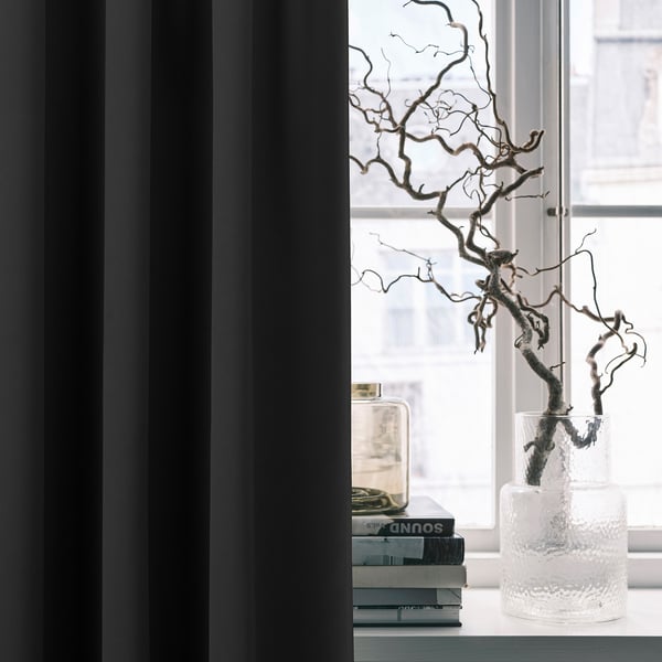 Décoration moderne de la chambre : des rideaux occultants MAJGULL encadrent une fenêtre avec un vase texturé contenant des branches séchées et des piles de livres.