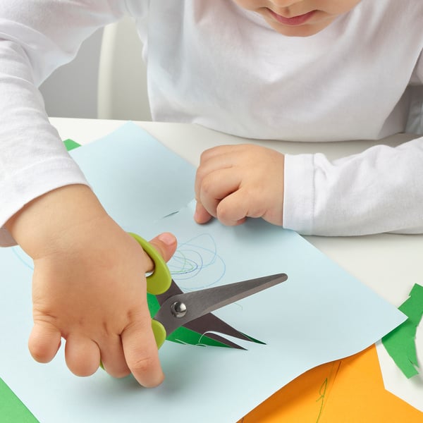 Enfant coupant du papier bleu clair avec des ciseaux à manche vert sur une table.