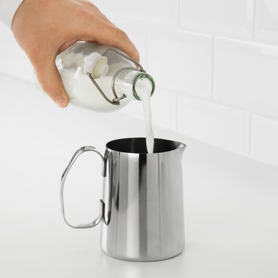Une main verse le lait d’une bouteille dans un pichet à mousse de lait MÅTTLIG en acier inoxydable, mettant en valeur sa conception facile à nettoyer et durable.