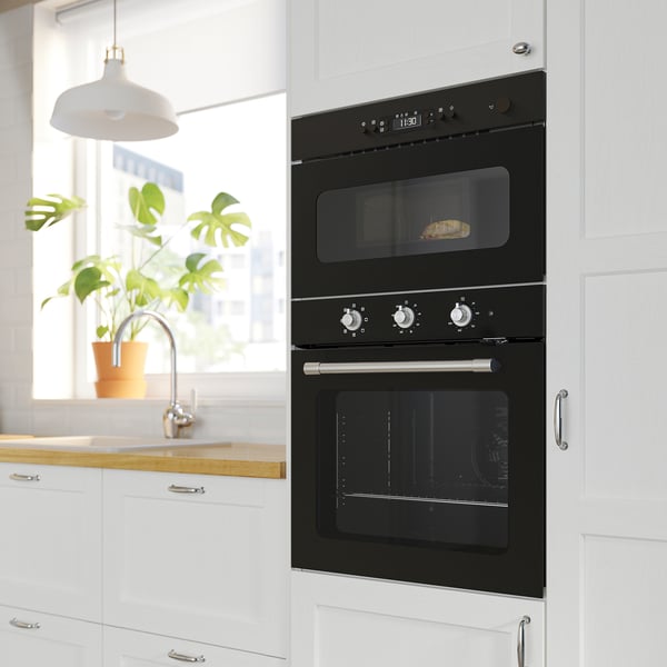 Cuisine moderne avec four noir et micro-ondes intégrés, armoires blanches, comptoir en bois, plante en pot et lampe suspendue.