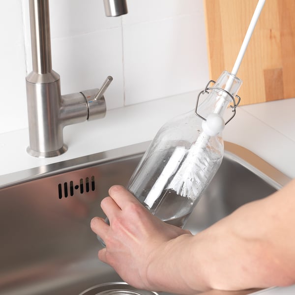 Personne tenant une bouteille transparente avec une brosse à long manche sous un robinet.