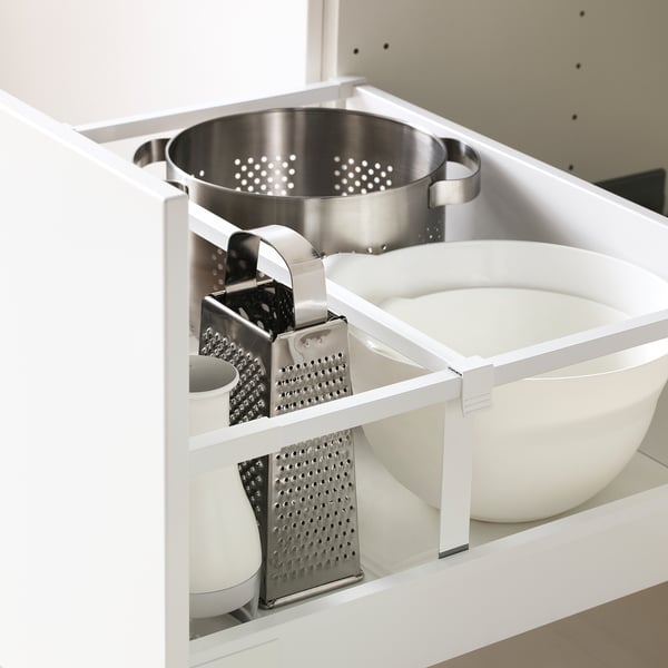 Armoire de cuisine blanche ouverte montrant une passoire en étain et des râpes soigneusement rangées sur une grille blanche.