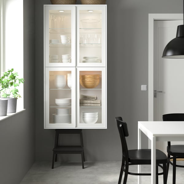 Scène de cuisine moderne avec une grande armoire blanche exposant de la verrerie et de la vaisselle. Cabinet flanqué de chaises en bois, près d’une fenêtre avec plante verte.
