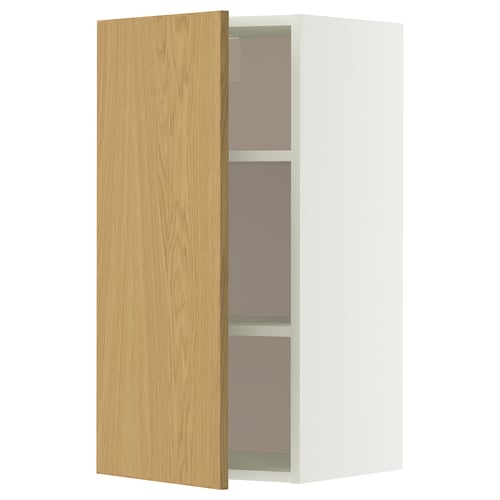 Une armoire murale METOD blanche avec une porte en chêne clair s’ouvre, révélant deux étagères en verre à l’intérieur.