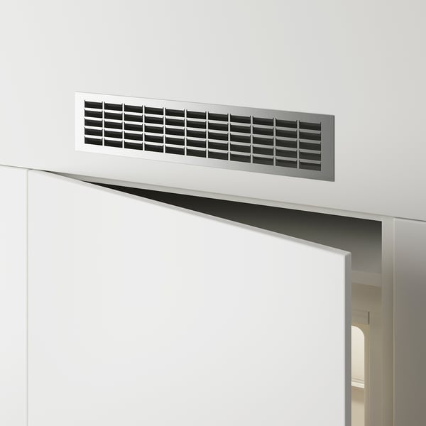 Grille de ventilation rectangulaire en métal montée sur un mur blanc au-dessus d’une porte, permettant la circulation de l’air.