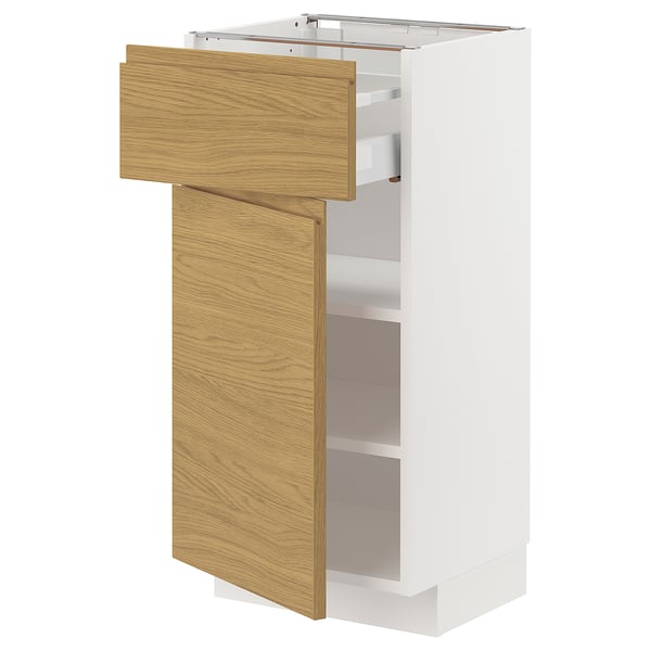 Un meuble de cuisine METOD blanc avec un tiroir et une porte en chêne clair, partiellement ouvert, révélant un espace de rangement à l’intérieur.