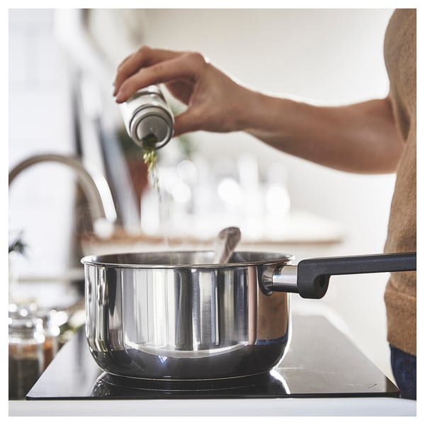 Personne assaisonnant des aliments dans une casserole en acier inoxydable sur une cuisinière. La casserole a une poignée noire et un couvercle en verre. La scène suggère une cuisine maison.