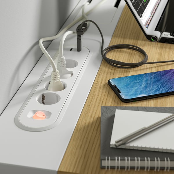 Bureau blanc avec prises de courant intégrées et un câble de chargeur enroulé. Un smartphone repose sur la surface en bois à côté d’un ordinateur portable.