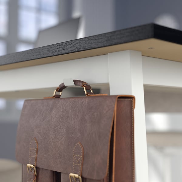 Un sac en cuir marron est suspendu à un crochet de tables hautes, avec un comptoir gris et une jambe blanche visibles.