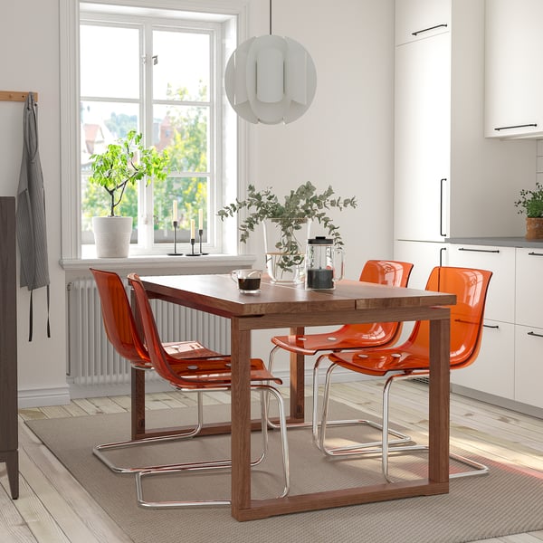 Salle à manger moderne avec des chaises orange et une table en bois, éclairée par un pendentif blanc, des plantes vertes et un tapis gris.