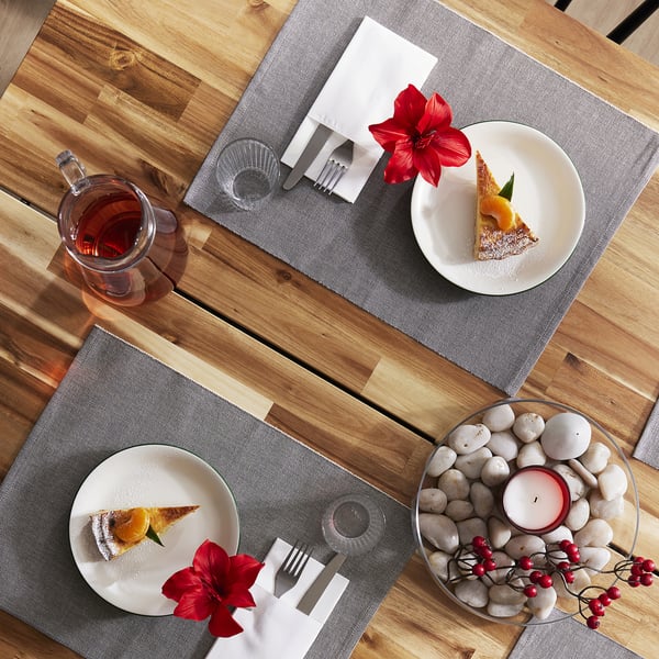 Deux tables en bois dressées avec des serviettes grises MOTTAGA, chacune contenant une assiette blanche surmontée d’un gâteau et d’une bougie, une fleur rouge et un verre transparent.