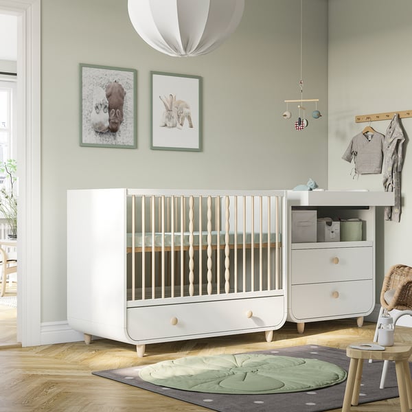 Chambre d’enfant moderne avec lit bébé blanc MYLLRA, impressions murales vertes, lampe suspendue et table à langer. Le lit bébé est grand, semblable à une commode.