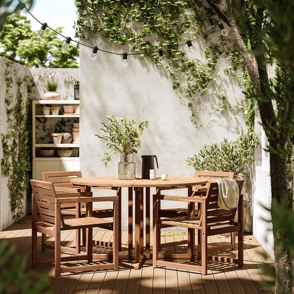 Ensemble de salle à manger extérieur avec 4 chaises et une table en bois, sous des guirlandes lumineuses et un mur blanc avec de la verdure.