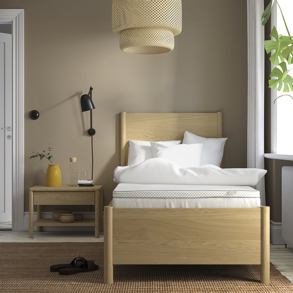 Chambre minimaliste avec lit en bois clair, literie blanche, table de chevet avec décoration, lampe suspendue, grande fenêtre et tapis.