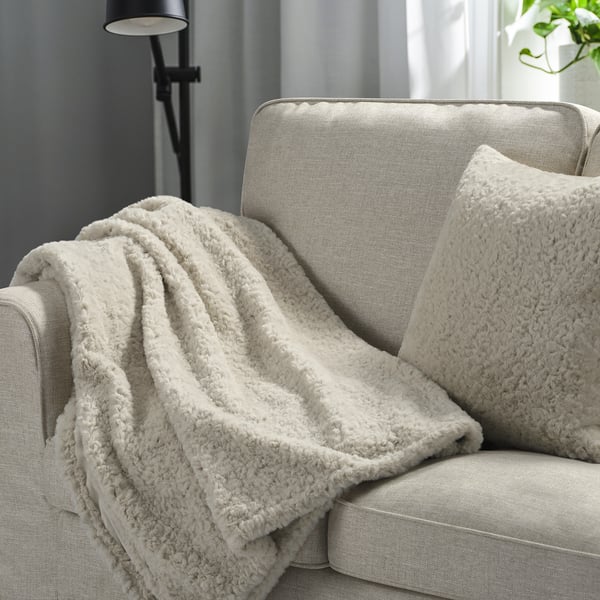 Couverture et oreiller beiges NÄTNÄVA sur canapé gris, créant une atmosphère chaleureuse.