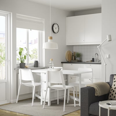 Cuisine moderne avec table NORDEN et chaises empilables. La table a des tiroirs, près de la fenêtre avec lampe suspendue. Armoires blanches, comptoir et tapis gris.
