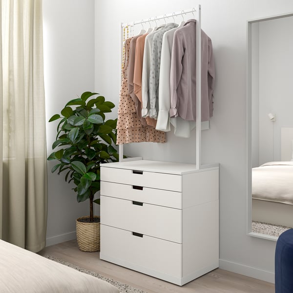 Chambre moderne avec coffre blanc IKEA NORDLI. Tringle à vêtements au-dessus, plante à côté et lit à proximité. Design épuré et minimaliste.