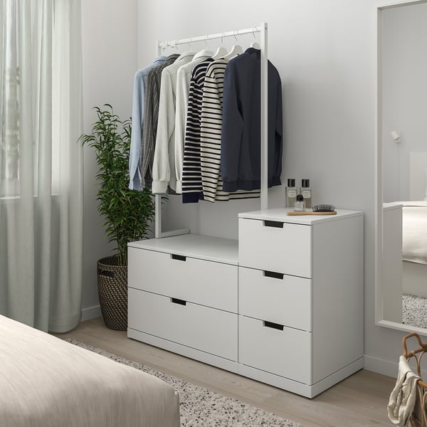 Commode NORDLI blanche moderne dans la chambre à côté du lit. Tringle à vêtements au-dessus, plante à côté.