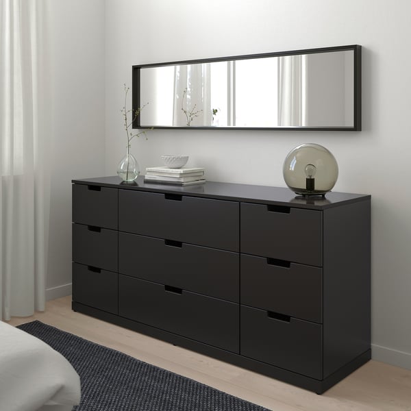 Coffre NORDLI noir moderne avec miroir, lampe, fleurs et livres. À côté du lit avec couverture blanche et tapis gris.