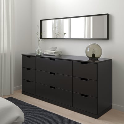 Coffre NORDLI noir moderne avec miroir, lampe, fleurs et livres. À côté du lit avec couverture blanche et tapis gris.