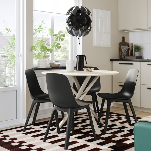 Table ronde NORRMJÖLE, chaises noires, tapis à damiers, table en mélamine, lumière suspendue, plantes.