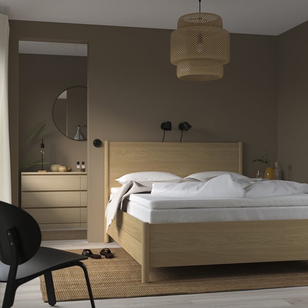 Chambre minimaliste : lit en bois clair, draps blancs, suspension tissée, murs sombres, chaise noire, tapis beige.