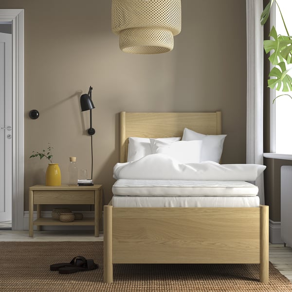 Lit minimaliste avec tête de lit en bois, draps blancs, suspension, table d’appoint avec lampe et vase jaune. Le lit est plus grand que la table d’appoint.