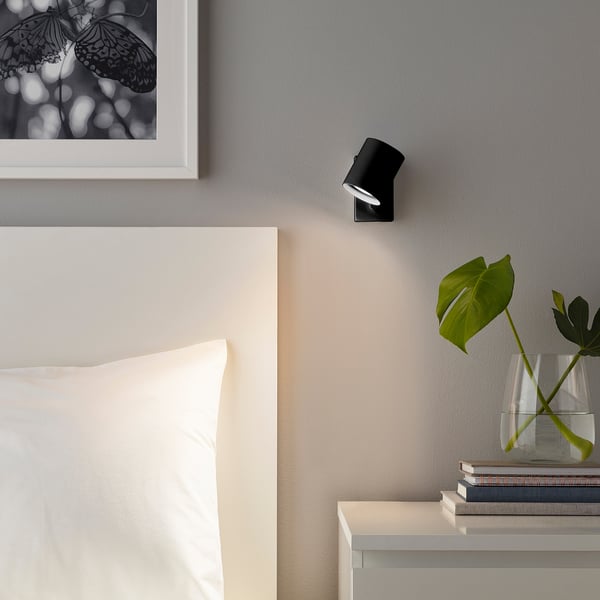 Applique murale NYMÅNE en noir près d’un lit, avec un vase et des œuvres d’art. Petit design élégant.