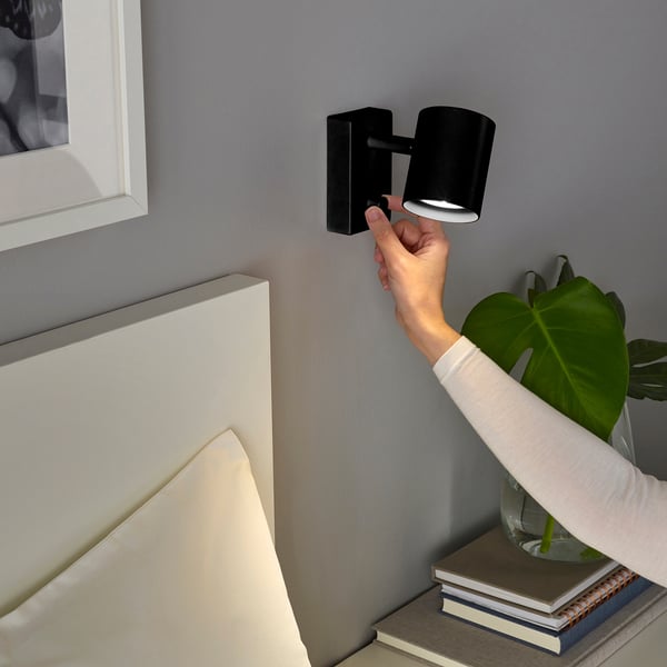 Personne ajustant une lampe murale noire près d’une tête de lit.