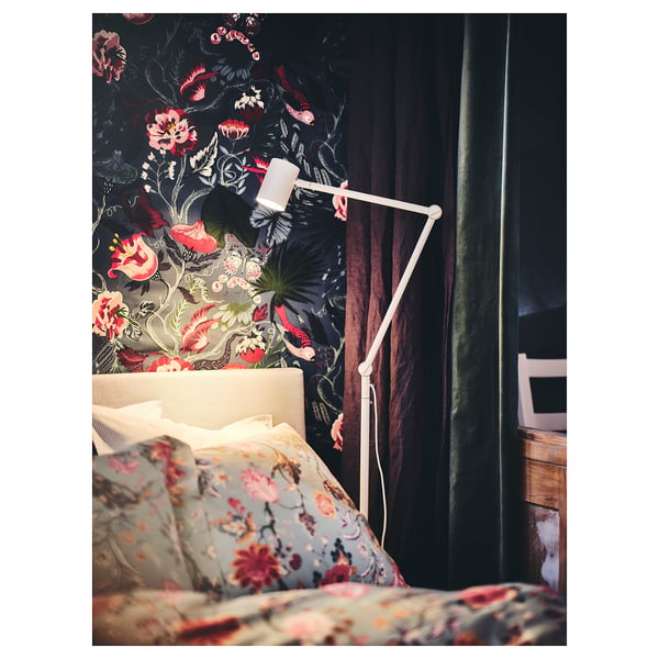 Une chambre avec un papier peint floral sombre et un lampadaire en métal blanc, illuminant des coussins floraux.