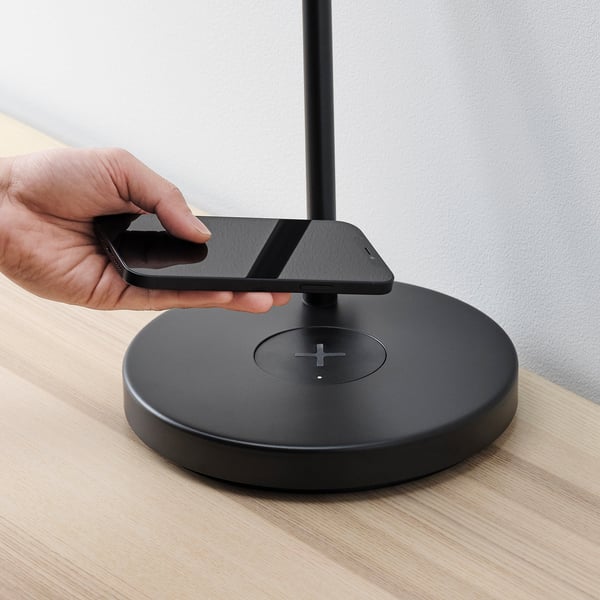La main pose le smartphone sur un socle de charge sans fil au sommet d’une base circulaire, à côté d’une lampe moderne sur la table.