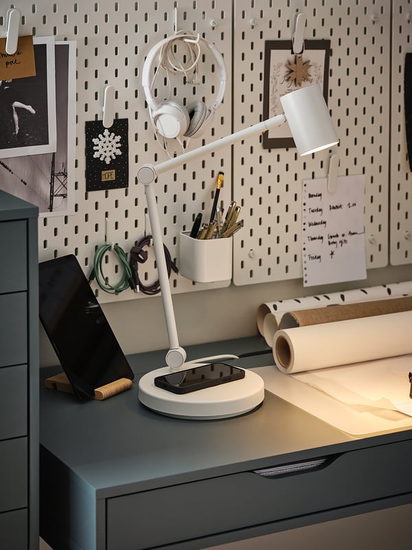 Espace de travail moderne avec lampe NYMÅNE, téléphone, porte-stylo, papier et panneau perforé avec notes et écouteurs.