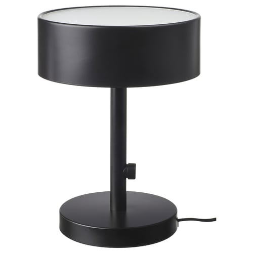 Une lampe de table NYMÅNE, noire, avec un abat-jour et une base cylindriques, en métal et en verre, fournissant une lumière douce et diffuse.