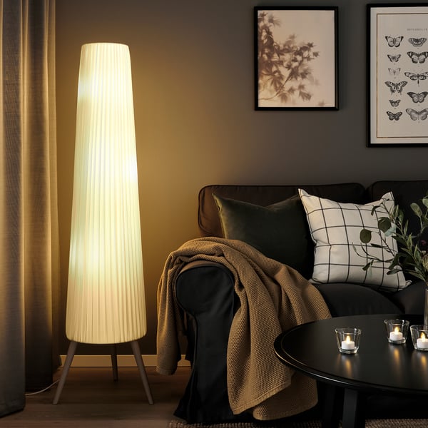 Lampadaire ÖKENSAND à côté du canapé, émettant une lumière douce, atteignant presque le plafond.